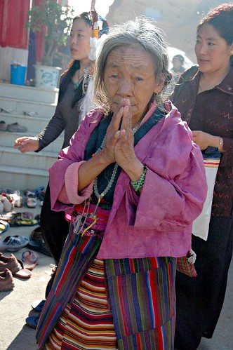 Elder Tibetan Woman, Bodhisattva Day, with her hands in prayer mudra, wearing traditional apron and wrap, malas, two Buddhist women, Tharlam Monastery, Boudha, Kathmandu, Nepal by Wonderlane