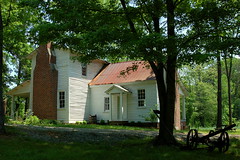 Photograph of a white house with wood siding, a brick chimney, and a red tile roof. It is built in the style of the late 18th or early 19th centuries. 