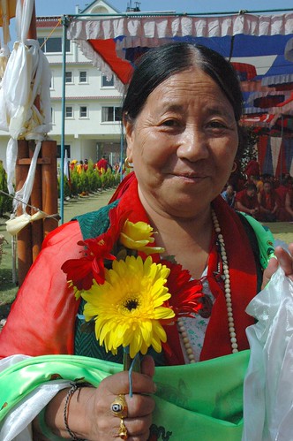 Tibetan Woman with flower and katag offerings, wearing a dzi stone ring, memorial wrist bracelet to Tibetan prisoners, and mala, Tharlam Monastery Courtyard, Boudha, Kathmandu, Nepal by Wonderlane