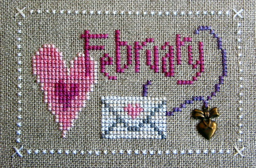 Cross stitch of February in pink thread on grey background with hearts and an envelope