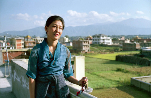 Tibetan woman, with a wistful expression - behind her is Tibet, wearing a blue chuba and a mala, holding car and house keys, on the roof of a house, overlooking the fields, Boudha, Nepal in 1990 by Wonderlane