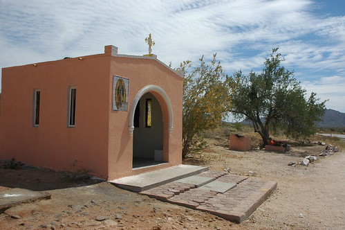 Our Lady of Guadalupe, cross, entrance, pink roadside shrine or chapel, a man sits on a concrete tank under a tree, mountains, clouds, Sonora Desert, northern Mexico, along the border, Highway 2 by Wonderlane