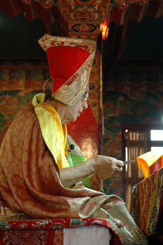 Guru Dagchen Rinpoche on his throne teaching at Sakya Lamdre, traditional silk robes, pecha, Sashu Hat (of the Sakya tradition), murals of the Buddha's life, Tharlam Monastery of Tibetan Buddhism, Boudha, Kathmandu, Nepal by Wonderlane