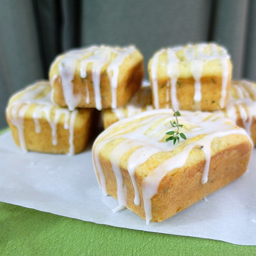 6 individual Lemon Thyme Tea Cakes staked two high on white paper. Glaze is dripping down to paper. A fresh thyme garnish is on top of front cake. 