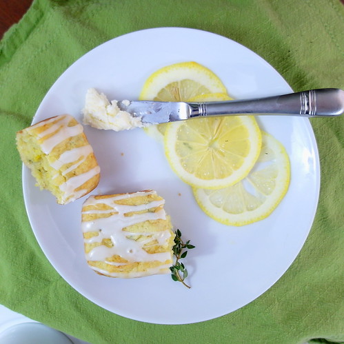 A single Lemon Thyme Tea Cakes, broken open, showing yellow crumbs. A silver knife has butter, ready for spreading. White plate has garnish of lemon slices and fresh thyme sprig.