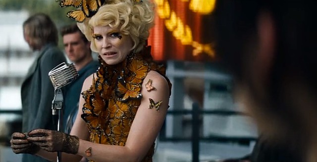 Effie Trinket, wearing a dress made of butterflys, seems upset while applauding in front of a microphone