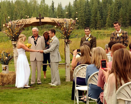 Jessie about to marry her beau, Chris, officiant, groomsmen in vintage clothing, photographer, audience with camera phones, country style, cross topping the white birch wedding arch, Fairbanks, Alaska, USA by Wonderlane