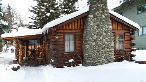 Daytime winter log cabin, doll on porch, wagon wheels, rock chimney, snow, South Addition, Anchorage, Alaska by Wonderlane
