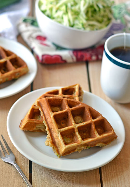 Waffles made with zucchini on two plates next to a cup of tea and a bowl of shredded zucchini