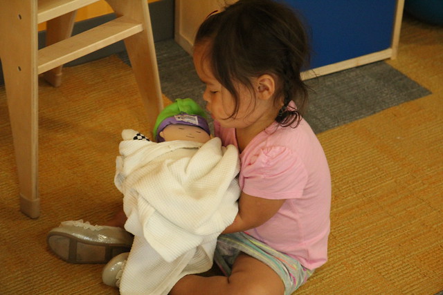 In the baby care area, holding a baby doll. Practicing to be a big sister?