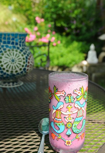 Boysenberry milkshake in a tumbler decorated with two mermaids, spoon, mesh patio table, blue glass candle lamp, white Buddha, Japanese lantern, pink carpet roses, A Garden for the Buddha, Seattle, Washington, USA by Wonderlane