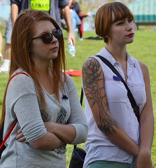 Two young girls, high school students, standing together listening, one with large tatoo of tree on her arm.