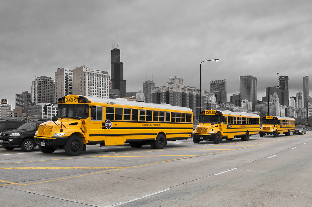 Three yellow school buses in a row traveling on the highway with a grey sky behind them