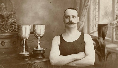 Man in a swimming costume standing with two trophies