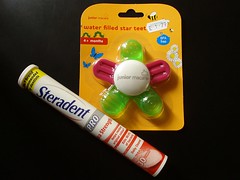 A tube of Steradent denture tablets and a cardboard/plastic blister pack containing a baby teether, both lying on a matte black background. The baby teether has “junior macare” printed in the middle of it, and five rounded nubs coming out from here; three green and filled with liquid and two pink and solid. The blister pack reads “water filled star teether” and there's a price sticker reading “£1.79”.