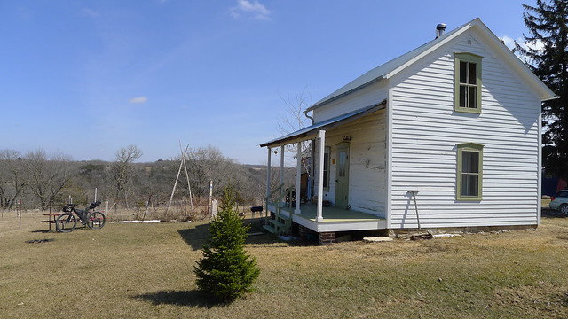 Left side view of an old, white farm house with a porch, on a grass plot, with bare trees in the distant background