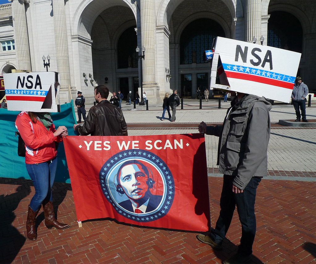 protesters holding a sign that says "yes we scan" and has a picture of Obama