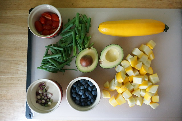 Ingredients laid out prettily on a cutting board: cut and uncut squash combine with ramekins of tomatoes, blueberries, and scallion slices to form a border around an angled pile of scallion greens and two halves of an avocado