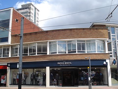 A large, modern-looking end-of-terrace shopfront with mannequins in the windows wearing suits and a sign reading “Moss Bros” above.  The first floor of the two-storey building has unusual zig-zag slanting windows.  Power cables for the tram network can be seen in the foreground.