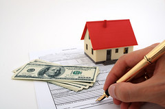 A hand holding a pen, signing a contract, with a house and some money in the background.