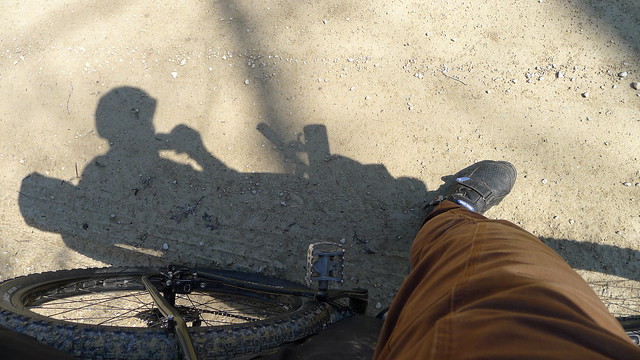 Downward view of a cyclist's leg, on the left side of a bike, on a gravelly ground
