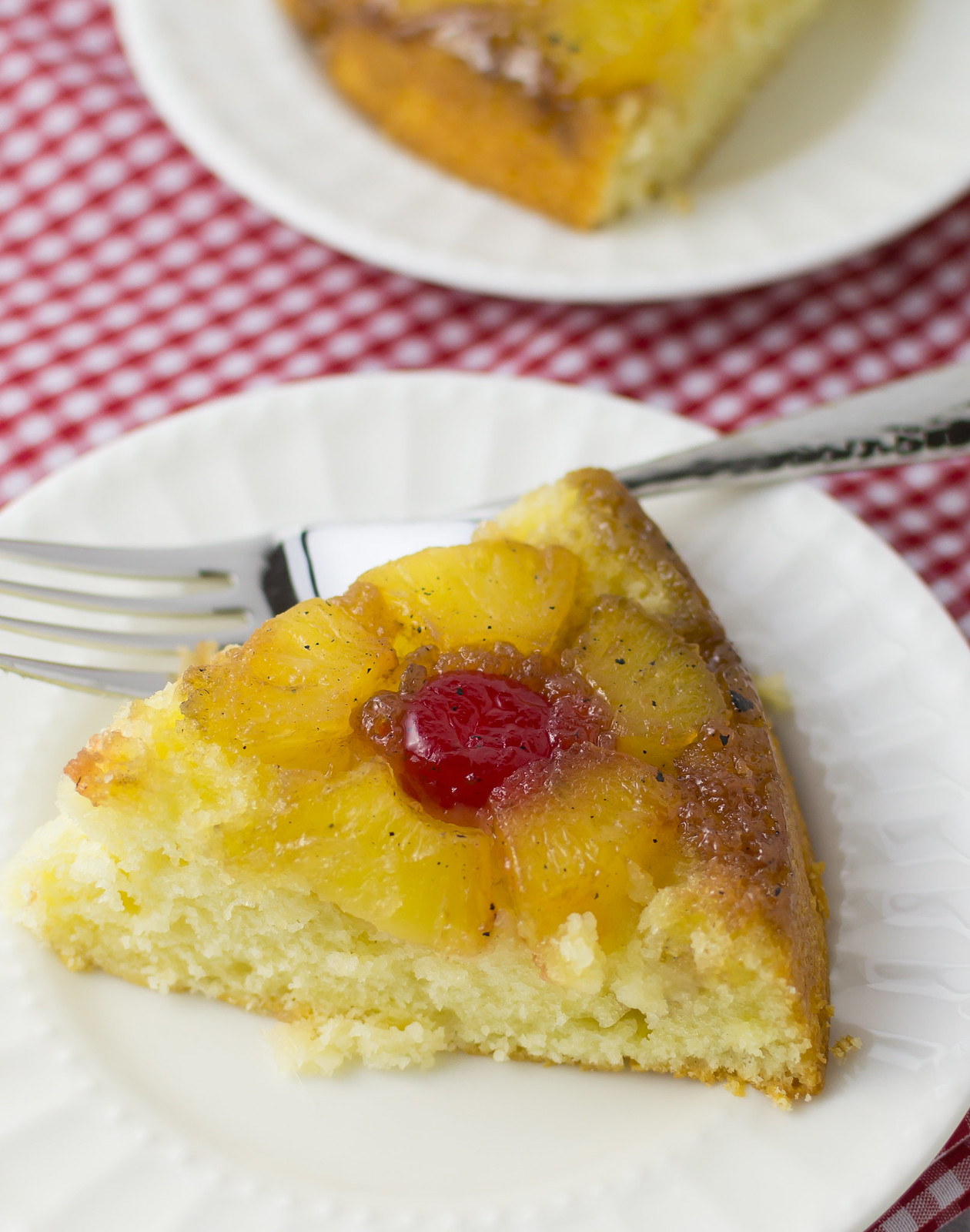 slice of prepared pineapple upside down cake on white plate with fork on red and white checkered napkin