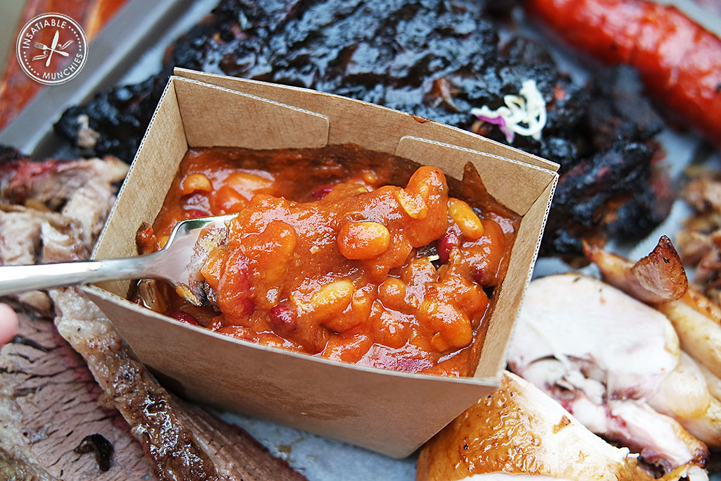 Thick rich baked beans sits in a brown, cardboard carton, and being scooped upwards with a fork.