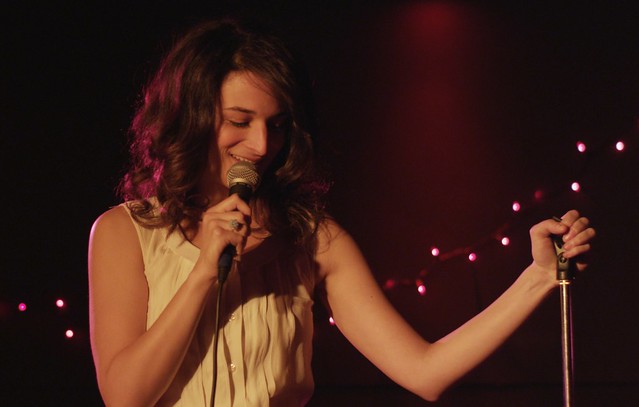 Jenny Slate stands on a stage, holding a microphone looking down