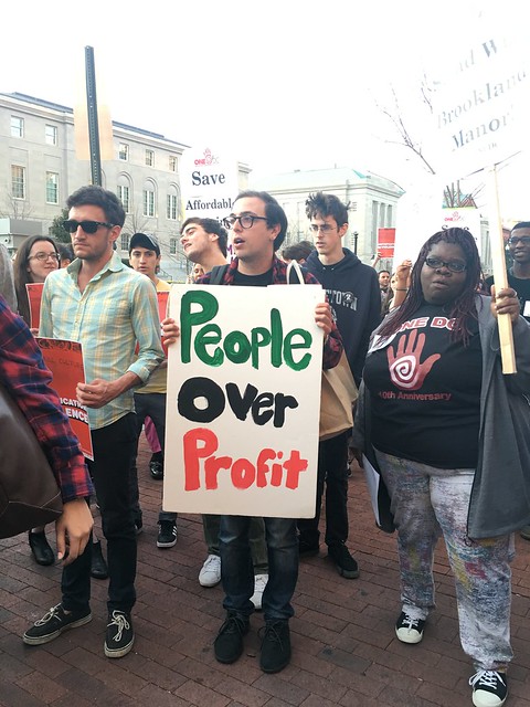 A crowd is gathered in front of the Wilson Building. A man holds a sign that reads, 