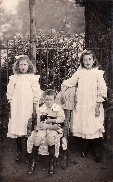 Old photograph of two girls with big bows in their hair and below the knee dresses with aprons and high-top boots standingon either side of a young man in short pants, black knee-his and books sitting in a wicker chair and holding a cat.